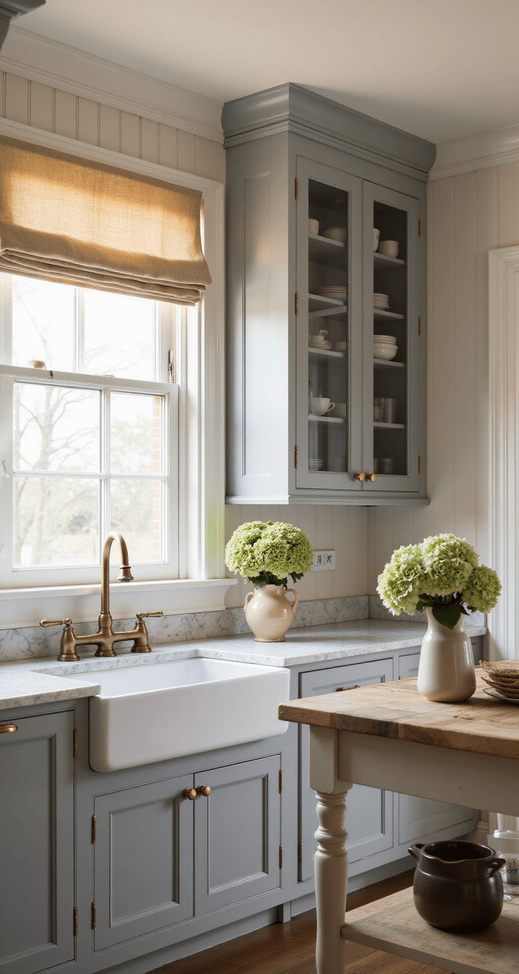 IKEA Kitchen Cabinets: The Ultimate Guide to Affordable, Stylish Storage Solutions An elegant BODBYN kitchen with soft gray raised-panel cabinets and brass hardware at golden hour, showcasing glass-front upper cabinets filled with vintage dishware, marble countertops, and a farmhouse sink. A butcher block island with turned legs complements the space, adorned with fresh hydrangeas in a ceramic vase. Natural linen window treatments filter warm light, capturing a timeless blend of traditional and modern styles.