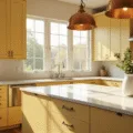 A spacious, sunlit modern kitchen featuring butter yellow Shaker cabinets, white quartz countertops, and vintage copper pendant lights, with natural light streaming through large windows.