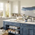 A bright and organized professional kitchen cabinet painting workspace featuring a mid-painting white cabinet door in Farrow & Ball Hague Blue, with high-end painting supplies, drop cloths on dark hardwood floors, and soft golden hour lighting enhancing the scene.