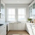 A bright, modern minimalist kitchen featuring floor-to-ceiling white cabinets with clear glass doors displaying neatly arranged white porcelain dinnerware and crystal glasses, complemented by white quartz countertops and stainless steel appliances, all bathed in morning sunlight.