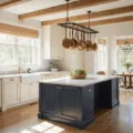 A sunlit farmhouse kitchen featuring cream Shaker cabinets, a navy blue island with marble countertops, and vintage copper pots, all illuminated by morning light streaming through mullioned windows.