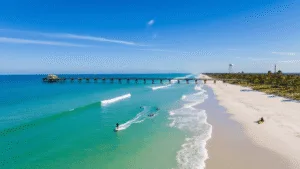 Why Cocoa Beach in January is Your Secret Winter Paradise "Surfer riding a wave on Cocoa Beach in January, with clear turquoise waters, empty white sand beach, Cocoa Beach Pier, palm trees, kayaking group, jumping dolphins, and Kennedy Space Center in the distance."