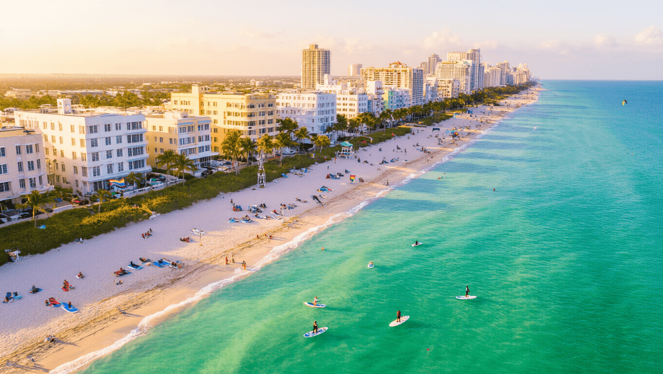 "Aerial view of South Beach Miami with Art Deco buildings, turquoise waters, white sandy beaches, beachgoers, paddle boarders, kite surfers, palm trees, lifeguard stands, and rainbow Pride flags in early morning sunlight."