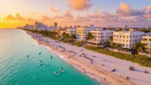 Miami Beach in October: Your Ultimate Tropical Escape Guide "Aerial view of Miami Beach at sunset in October, featuring Art Deco buildings with Halloween decorations, palm trees, turquoise waters, white sand beaches, beachgoers, paddleboarders, jet skiers, rooftop bars and colorful umbrellas"
