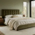 A serene modern bedroom with olive velvet tufted headboard, layered cream linen bedding, and floating walnut nightstands, bathed in warm golden hour light streaming through floor-to-ceiling windows, featuring bleached oak herringbone flooring and a textured wool area rug.
