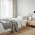 A serene Scandinavian guest bedroom featuring a twin bed with crisp white linens, soft gray throws, light blonde wood nightstand, a ceramic lamp, and natural light filtering through sheer linen curtains, set on a light wooden floor with a jute rug and knitted cream pouf.