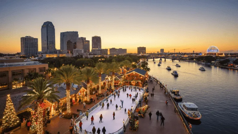 Why Tampa in December Might Just Be Your Perfect Winter Escape (Without the Freezing Cold!) "Aerial view of downtown Tampa during December sunset with Christmas decorations, lit Tampa Riverwalk, Curtis Hixon Waterfront Park's Winter Village, decorated boats in the Hillsborough River, and Busch Gardens' lights in the distance."