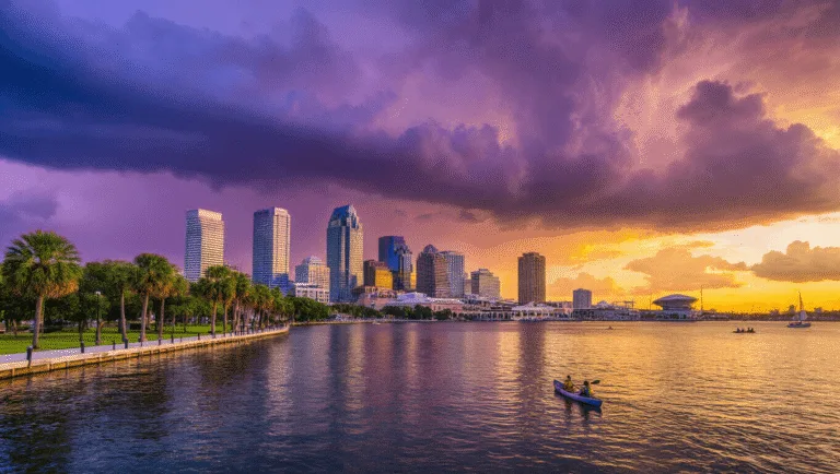 Why Tampa in September Will Blow Your Mind: A Local's Insider Guide "Tampa cityscape at sunset with skyline reflected in Tampa Bay, palm trees, kayakers, sailboat, Tampa Museum of Art, Raymond James Stadium, lush parks along the shoreline, and storm clouds building in the distance"