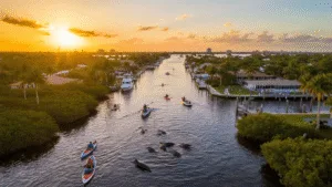 Fun Things to Do in Cape Coral: Your Ultimate Guide to Florida's Hidden Adventure Paradise "Aerial view of Cape Coral's canal system at sunset with various activities including kayaking, dolphins swimming, and owls nesting; Yacht Club Park and pier visible in the distance."