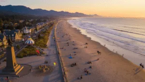 The Ultimate Seaside Adventure: Unleash Your Coastal Playground! "Aerial view of Seaside, Oregon's coastline at sunset with families, surfers, and horseback riders, featuring the promenade, Lewis & Clark monument, and Captain Kid Amusement Park."