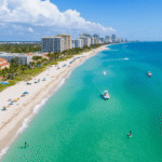 "Aerial view of Fort Lauderdale's coastline with turquoise waters, sandy beaches, palm trees, Las Olas Boulevard, boats on the Intracoastal Waterway, paddleboarders, and people enjoying a warm 76°F day."