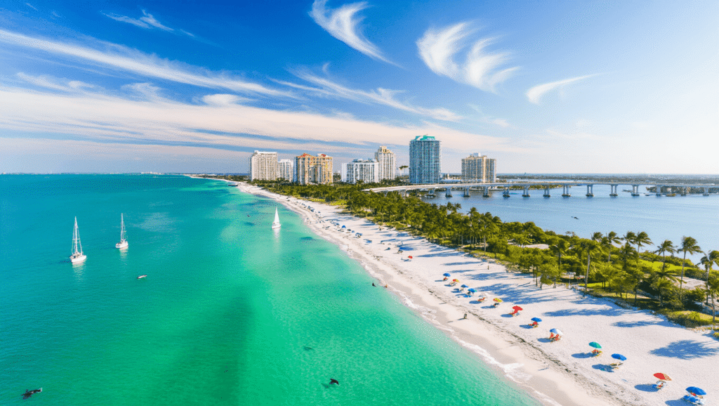 Sarasota in April: Your Ultimate Gulf Coast Paradise Revealed "Aerial view of Sarasota's coastline and Siesta Key beaches with turquoise waters, sailboats, palm trees, beach umbrellas, the John Ringling Causeway Bridge, and the Marie Selby Botanical Gardens under an April mid-afternoon sun."